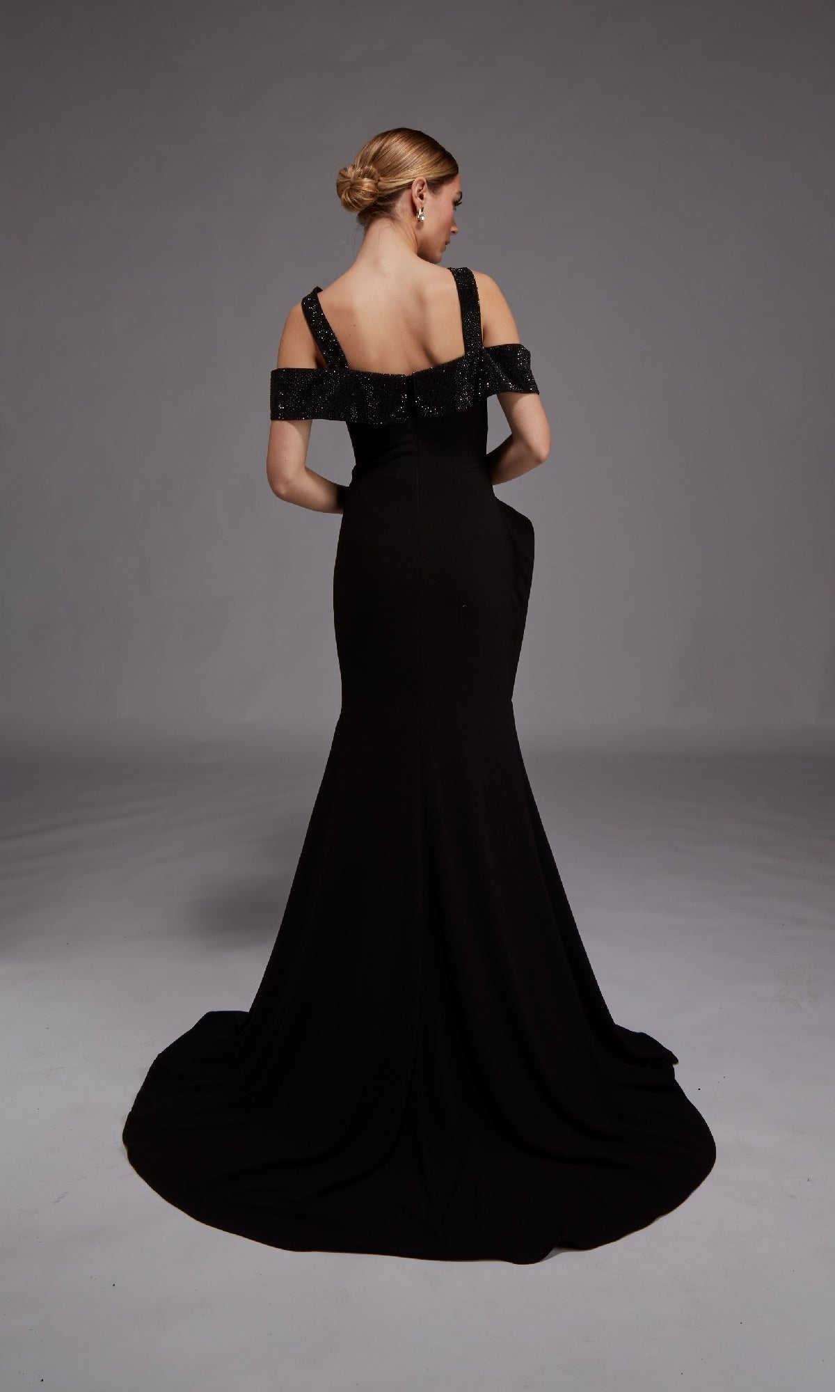 A woman faces away, wearing the Alyce 27878 long formal dress in black with a cold shoulder design, a fitted bodice, and a dramatic train, set against a plain gray background.